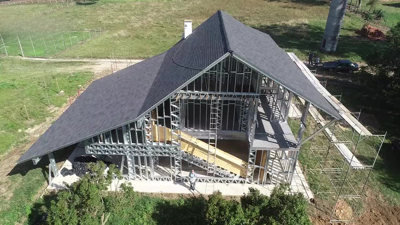 Casa em light steel frame em obra, vista de drone: a estrutura de perfis de aço
            à vista debaixo da cobertura já telhada, rodeada de relva e árvores.